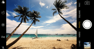 Regra dos terços: Vista panorâmica de uma praia tropical com céu azul claro, várias palmeiras inclinadas sobre uma faixa de areia onde pessoas estão tomando sol e nadando no oceano. Um veleiro aparece ao longe. A interface da câmera do smartphone exibe opções como HDR, flash e modos de captura, indicando a possibilidade de fotografias de alta qualidade durante as férias.