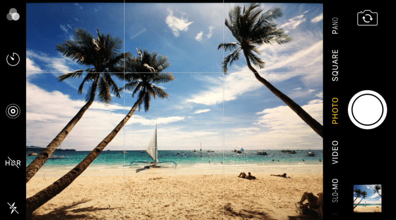 Regra dos terços: Vista panorâmica de uma praia tropical com céu azul claro, várias palmeiras inclinadas sobre uma faixa de areia onde pessoas estão tomando sol e nadando no oceano. Um veleiro aparece ao longe. A interface da câmera do smartphone exibe opções como HDR, flash e modos de captura, indicando a possibilidade de fotografias de alta qualidade durante as férias.